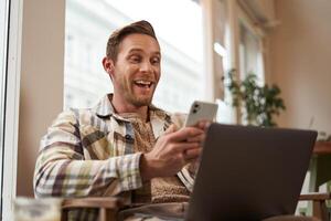 image de homme dans café, café magasin visiteur séance dans chaise avec portable et téléphone intelligent, à la recherche surpris et excité à mobile téléphone filtrer, étonné par gros nouvelles, promo offre sur app photo