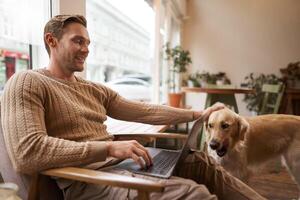 Beau Jeune homme travail dans café avec une chien, séance sur chaise et en utilisant ordinateur portable, caresse le sien d'or retriever dans respectueux des animaux cotravail espace photo