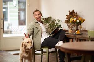 portrait de content souriant Jeune homme, café visiteur, séance dans café magasin avec le sien chien, caresse d'or retriever, en portant numérique tablette, en train de lire nouvelles ou navigation sur l'Internet photo