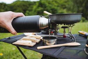 l'eau est versé de une thermos dans une tasse, le concept de une touristique vacances dans nature, une camp cuisine cuisson, chaud thé, camping ustensiles. photo