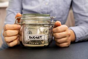 méconnaissable femme en portant économie argent dans verre pot rempli avec dollars billets de banque. budget transcription dans de face de pot. gérant personnel finances supplémentaire le revenu pour futur insécurité Contexte photo