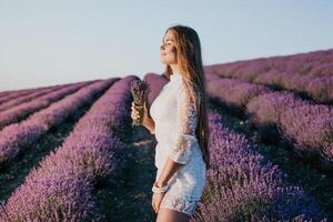 femme lavande champ. content insouciant femme dans une blanc robe en marchant dans une lavande champ et odeur une lavande bouquet sur le coucher du soleil. idéal pour chaud et inspirant concepts dans esprit d'aventure et Voyage. photo