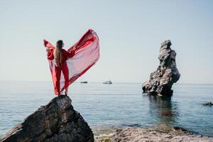 femme Voyage mer. Jeune content femme dans une longue rouge robe posant sur une plage près le mer sur Contexte de volcanique rochers, comme dans Islande, partage Voyage aventure périple photo