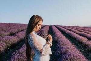 femme lavande champ. content insouciant femme dans une blanc robe en marchant dans une lavande champ et odeur une lavande bouquet sur le coucher du soleil. idéal pour chaud et inspirant concepts dans esprit d'aventure et Voyage. photo