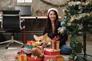 portrait d'une travailleuse asiatique avec un chapeau rouge et son chien regardant la caméra près de la décoration de l'arbre de Noël et des cadeaux, préparation du festival du bonheur et célébration de la fête du nouvel an sur un lieu de travail. photo