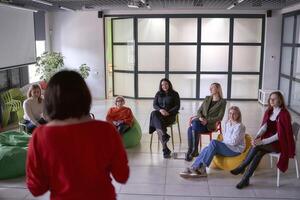 une femme fait du une discours sur organiser, collègues Ecoutez à sa discours et asseoir sur chaises et poufs dans le salle photo