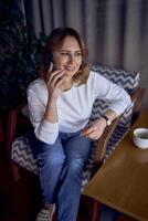 une femme dans une chandail et jeans est parlant sur le téléphone dans une magnifique café avec verdure et lumière panneaux dans le Contexte photo