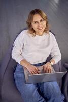 femme sur une bouffée chaise travail sur sa portable dans cotravail avec moderne intérieur conception, neutre tons et vert plante photo