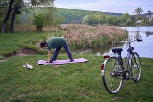 vieux femme Faire des exercices dans le Matin sur le rivière banque, une vélo et une tapis de yoga suivant à sa photo