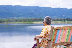 retour vue de une Sénior femme à la recherche à le Lac et montagnes tandis que séance sur une en bois chaise côté de le lac. concept de vieilli gens et relaxation photo