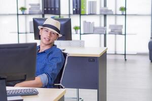 Jeune homme d'affaire porte une bleu chemise et chapeau séance sur une chaise dans le bureau. il est souriant et content à du repos après travail et vacances bientôt. concept de affaires et relaxation photo