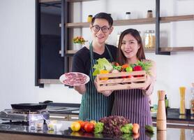 asiatique Jeune couple est en portant une boîte de des fruits et des légumes biologique et une plat de porc à préparer le steak dans le cuisine. concept de couple et santé nourriture biologique photo