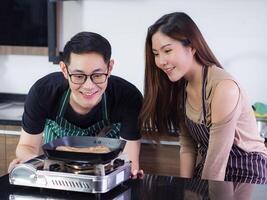 asiatique Jeune couple portant portion à faire une steak à le cuisine dans leur Accueil gril le steak sur une petit gaz le fourneau dans le cuisine. femme et homme sourire et Regardez steak dans une poêle. nourriture concept photo