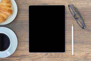 Haut vue de une bureau avec une tablette, crayon, lunettes, une tasse de café, et croissant sur une assiette mis sur une en bois tableau. travail de maison. espace pour texte photo