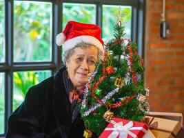 Sénior asiatique femme en portant une cadeau boîte et séance près une Noël arbre à maison. concept de vieilli gens et Festival photo