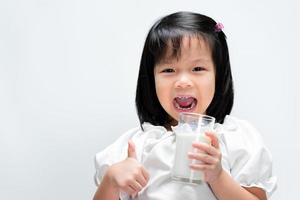 Une fillette de 4 ans boit du lait avec un verre. petit enfant a levé le pouce montrant que c'était bon. fond blanc isolé. photo