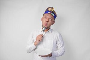 réfléchi Jeune balinais homme dans blanc chemise et traditionnel coiffure en portant un vide assiette avec copie espace et cuillère tandis que en pensant quoi nourriture à manger photo