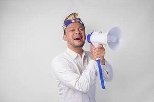 énergique balinais homme dans blanc chemise et traditionnel coiffure en criant à mégaphone tandis que élevage serré poing, isolé par blanc Contexte photo