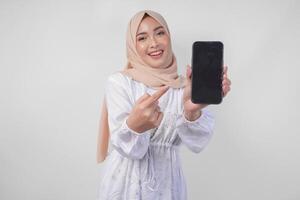 portrait de Jeune asiatique musulman femme portant blanc robe et hijab souriant Heureusement, montrer du doigt à le Vide écran copie espace sur sa téléphone et montrant écran à le caméra photo