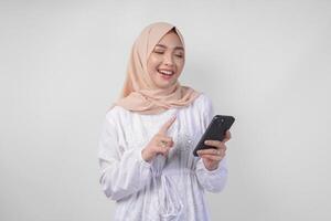 excité asiatique musulman femme portant blanc robe et hijab en portant sa téléphone intelligent tandis que souriant gaiement, isolé par blanc Contexte. ramadhan et eid mubarak concept photo