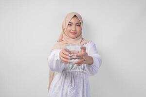 souriant asiatique musulman femme dans blanc robe et hijab en portant et en présentant une tasse de Frais eau, permanent plus de isolé blanc Contexte photo