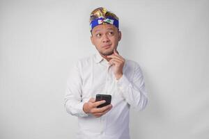 sérieux Jeune balinais homme dans blanc chemise et traditionnel coiffure en portant téléphone intelligent tandis que en pensant difficile photo