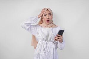 Jeune asiatique musulman femme portant blanc robe et hijab montrant sous le choc expression sur sa visage tandis que en portant téléphone intelligent, surpris après en train de lire nouvelles ou potins, isolé par blanc Contexte photo