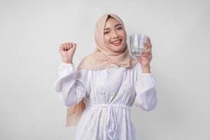 de bonne humeur asiatique musulman femme portant blanc robe et hijab en buvant une verre de l'eau avec une content réussi expression et geste, sentiment soulagé après jeûne, isolé par blanc Contexte. photo