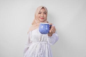 souriant asiatique musulman femme dans blanc robe et hijab en portant et en présentant l'eau ou boisson dans une bleu tasse, permanent plus de isolé blanc Contexte photo