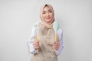 excité Jeune asiatique musulman femme dans une voile hijab et crème tablier souriant à le caméra tandis que en portant spatule et cuisine cuisine ustensiles photo