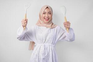 portrait de une content asiatique musulman femme dans voile hijab en portant spatule et cuisine cuisine ustensiles tandis que souriant gaiement. Ramadan concept photo