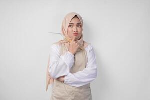 réfléchi Jeune asiatique musulman femme dans hijab et crème tablier en portant spatule et cuisine cuisine ustensiles, en pensant difficile quoi nourriture menu à cuisinier photo