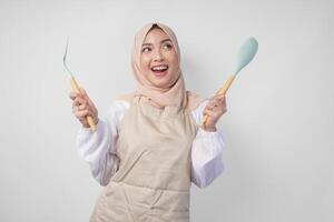 excité Jeune asiatique musulman femme dans une voile hijab et crème tablier souriant à le caméra tandis que en portant spatule et cuisine cuisine ustensiles photo