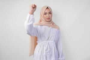 Jeune asiatique musulman femme portant blanc robe et hijab avec sérieux expression élevage une serré poing geste plus de isolé blanc Contexte photo