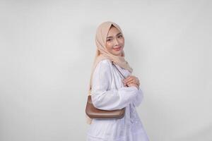 élégant Jeune asiatique musulman femme portant blanc robe et hijab avec marron fronde sac souriant Heureusement plus de isolé blanc Contexte. ramadhan mode concept photo