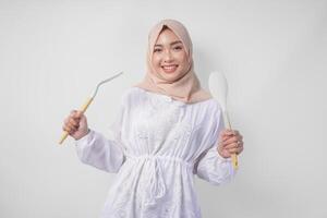 portrait de une content asiatique musulman femme dans voile hijab en portant spatule et cuisine cuisine ustensiles tandis que souriant gaiement. Ramadan concept photo