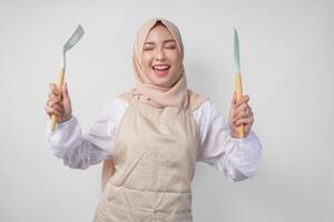 excité Jeune asiatique musulman femme dans une voile hijab et crème tablier souriant à le caméra tandis que en portant spatule et cuisine cuisine ustensiles photo