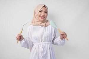 portrait de une content asiatique musulman femme dans voile hijab en portant spatule et cuisine cuisine ustensiles tandis que souriant gaiement. Ramadan concept photo