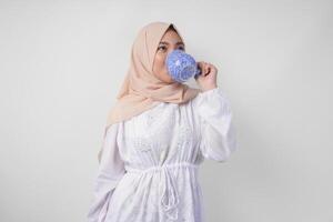 soif Jeune asiatique musulman femme dans blanc robe et crème voile hijab en buvant Frais minéral l'eau de une bleu tasse après jeûne. Ramadan concept photo
