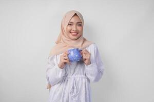souriant asiatique musulman femme dans blanc robe et hijab en portant et en présentant l'eau ou boisson dans une bleu tasse, permanent plus de isolé blanc Contexte photo
