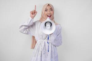 de bonne humeur asiatique femme portant hijab et blanc robe montrer du doigt en haut à le copie espace au dessus tandis que en criant à le mégaphone, permanent plus de isolé blanc Contexte photo