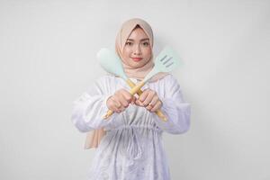 mécontent asiatique femme dans voile hijab Faire une refus ou rejet signe, en disant Non, demander à Arrêtez geste en utilisant spatule et cuisine ustensiles photo