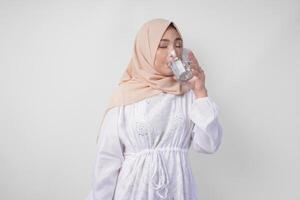 soif Jeune asiatique musulman femme dans blanc robe et crème voile hijab en buvant Frais minéral l'eau après jeûne. Ramadan concept photo