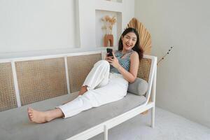 portrait de content asiatique femme souriant et en portant sa téléphone intelligent tandis que pose vers le bas sur une canapé dans villa pour été vacances photo