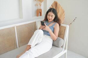 portrait de content asiatique femme souriant et en portant sa téléphone intelligent tandis que pose vers le bas sur une canapé dans villa pour été vacances photo