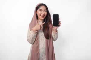 excité Jeune asiatique musulman femme dans foulard voile hijab montrer du doigt à le copie espace à téléphone intelligent Vide filtrer, isolé sur blanc Contexte studio. Ramadan et eid mubarak concept. photo