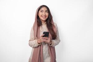 réfléchi asiatique musulman femme en portant une téléphone intelligent tandis que se demander et en pensant avec copie espace au-dessus de, isolé par blanc Contexte photo