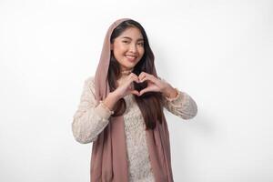 une content Jeune asiatique musulman femme portant voile hijab fabrication l'amour cœur forme en utilisant mains sur isolé blanc Contexte. Ramadan et eid mubarak concept photo