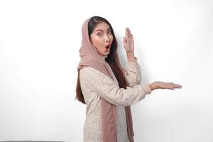 surpris asiatique musulman femme portant foulard voile hijab à la recherche choc tandis que montrer du doigt à copie espace à côté de son, permanent sur isolé blanc Contexte photo