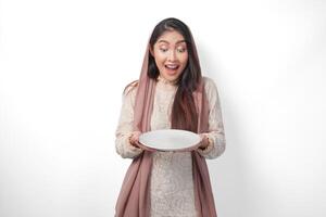 surpris asiatique musulman femme dans foulard voile hijab en portant assiette avec copie espace à l'intérieur, isolé blanc Contexte photo